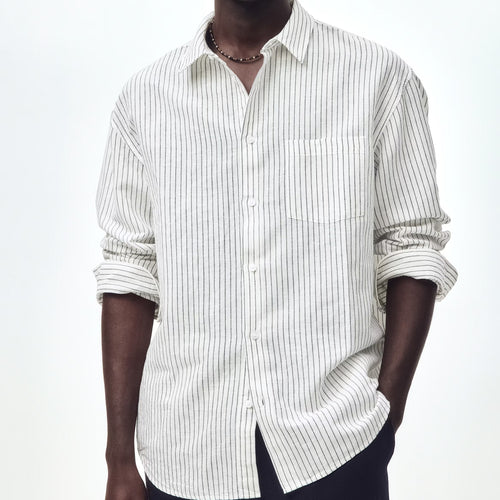 A man wearing a white long-sleeve button-up shirt with thin vertical navy stripes, styled with dark trousers, sunglasses, and a necklace against a plain white background.