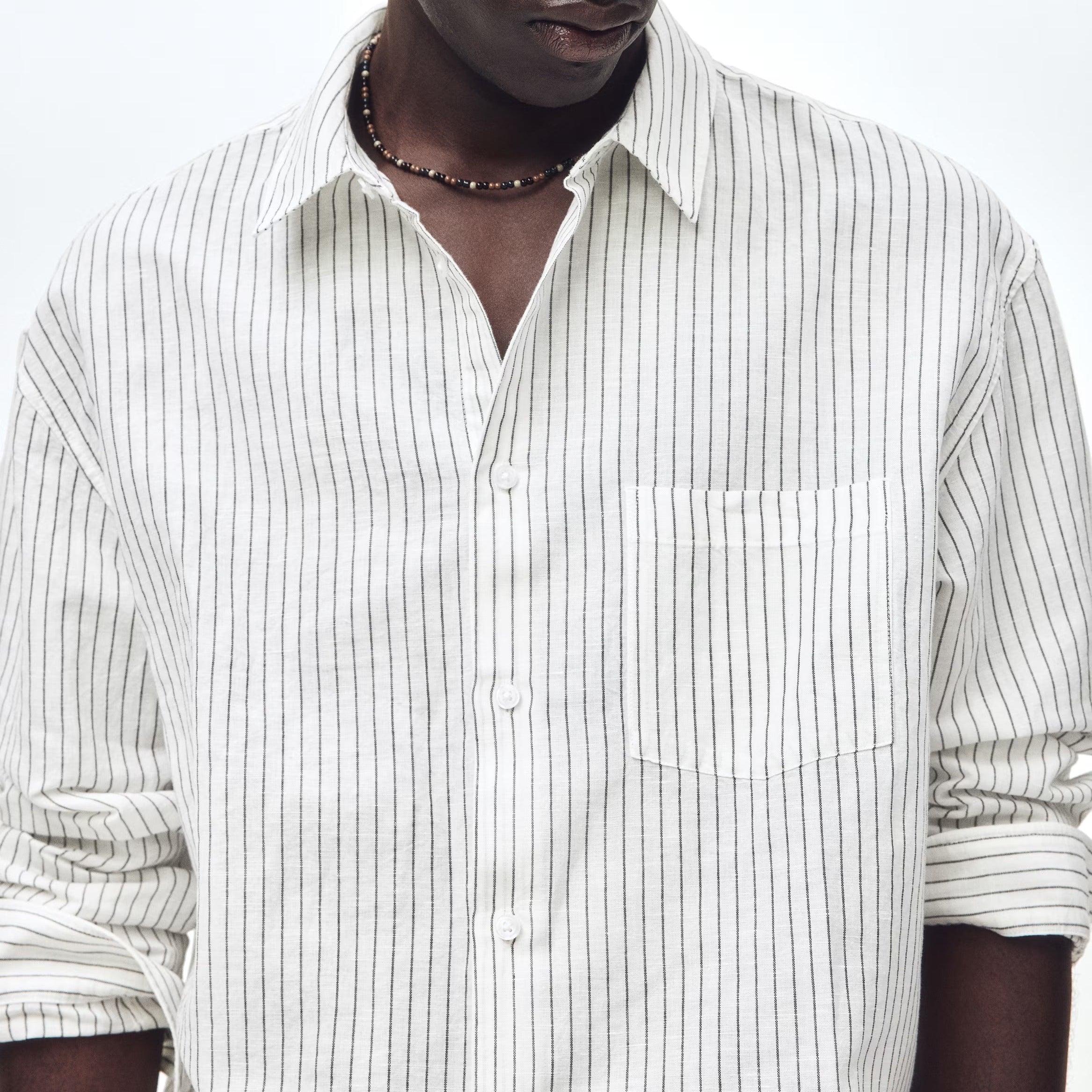 A man wearing a white long-sleeve button-up shirt with thin vertical black pinstripes, styled with a dark beaded necklace, against a plain white studio background.