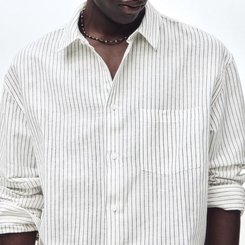 A man wearing a white long-sleeve button-up shirt with thin vertical black pinstripes, styled with a dark beaded necklace, against a plain white studio background.