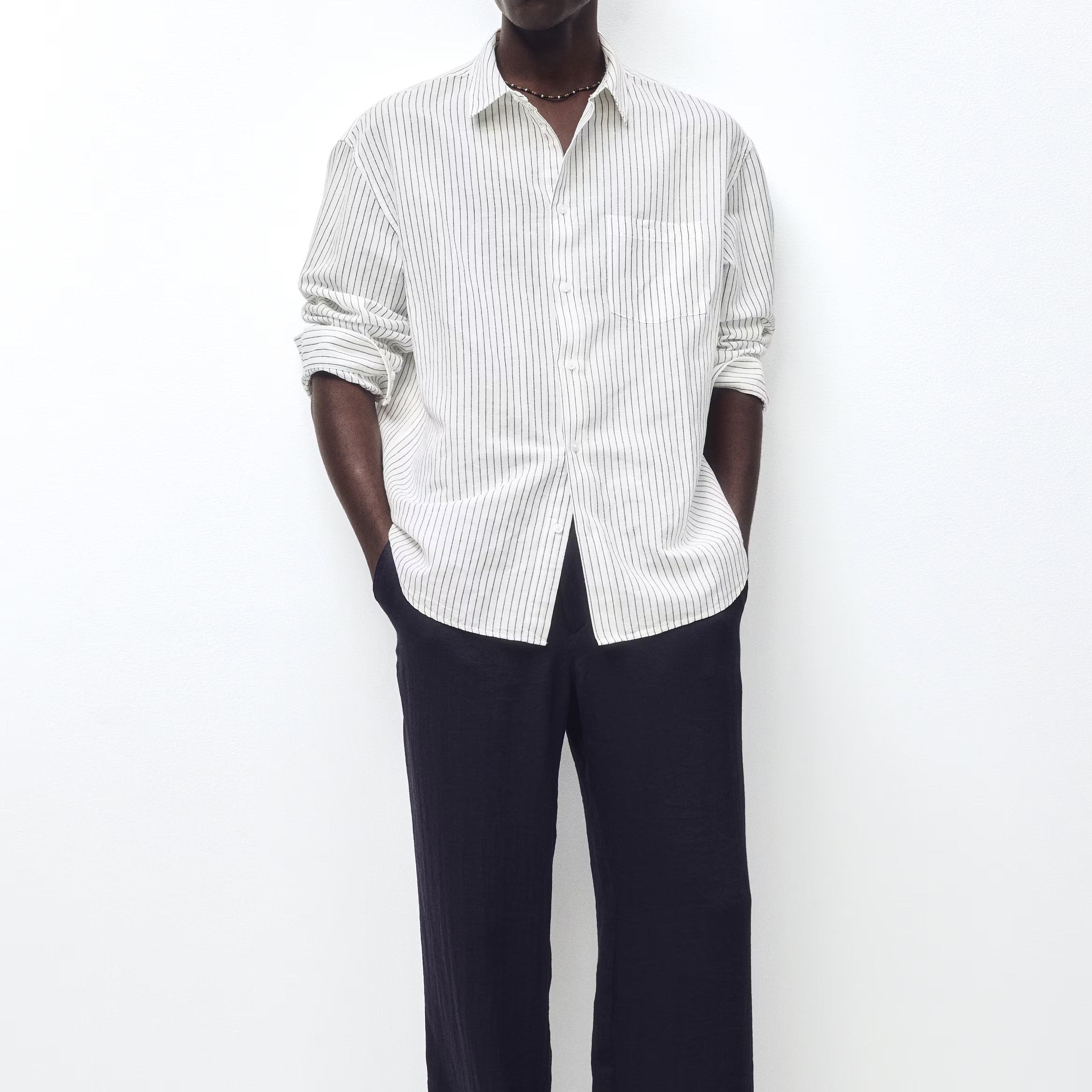 A man wearing a white long-sleeve button-down shirt with thin vertical dark stripes, paired with dark wide-leg trousers and sunglasses, standing against a plain white wall.