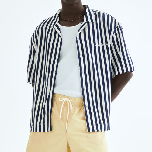A man modeling a short-sleeve button-up shirt with navy and white vertical stripes, worn open over a white t-shirt and paired with light yellow drawstring shorts, against a plain white studio background.