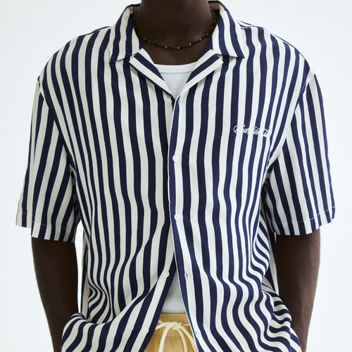 A man wearing a navy and white vertically striped short-sleeve button-up shirt with a small white embroidered logo on the chest, paired with light yellow drawstring shorts against a plain white background.