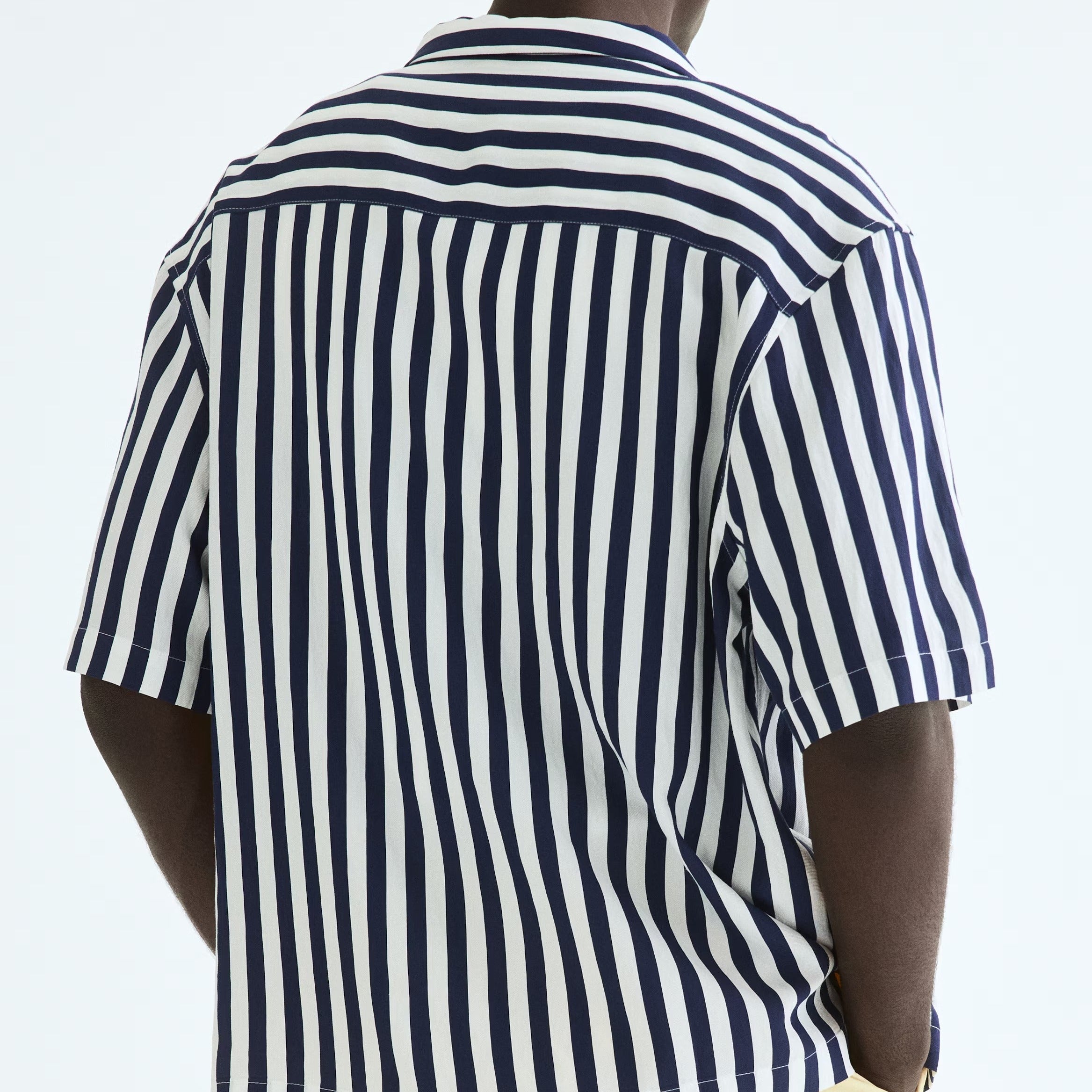 A man viewed from behind wearing a short-sleeve button-up shirt with vertical navy and white stripes, paired with light yellow pants, against a plain white background.