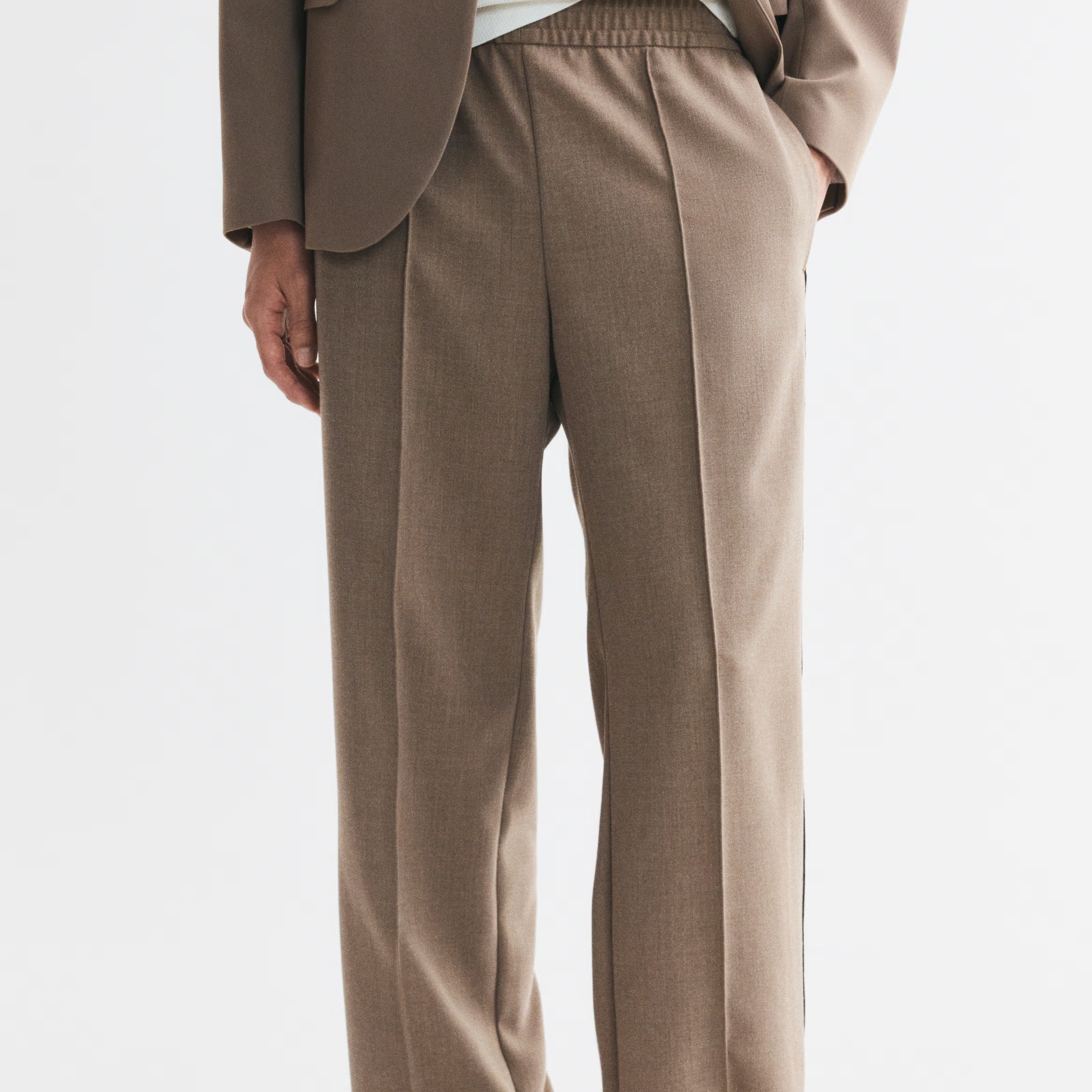 A person wearing loose, wide-leg taupe trousers with an elastic waistband, styled with a white top, brown blazer, and black sandals against a plain light gray studio background.