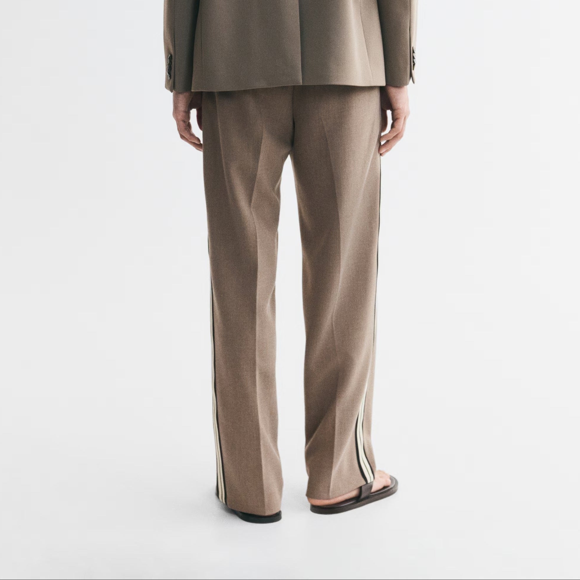 A man viewed from behind wearing a matching taupe-brown suit, featuring a structured blazer and wide-leg trousers with thin light-colored side stripes, set against a plain white studio background.