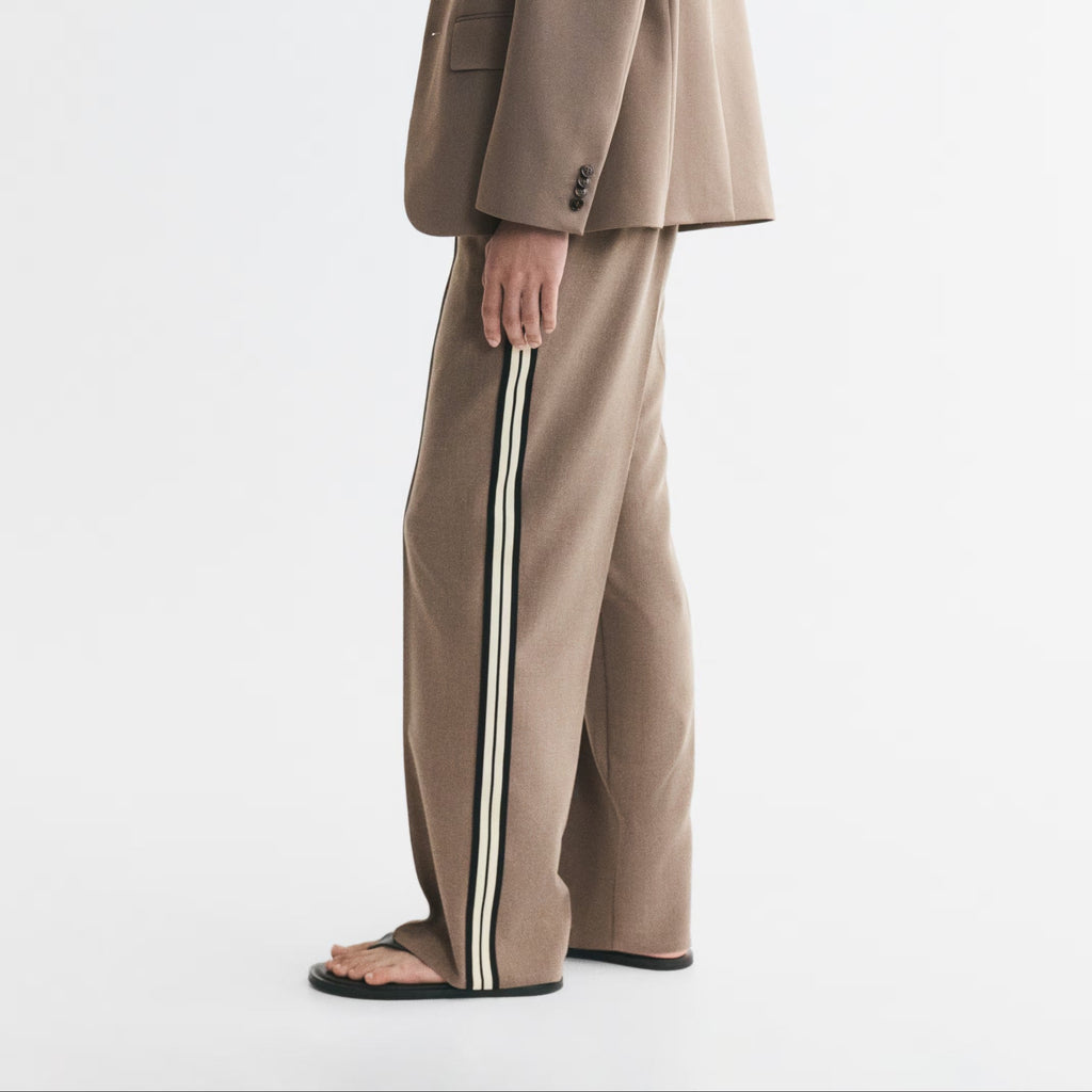 A man wearing a beige two-piece suit with wide-leg trousers featuring a double white stripe down the side, standing in profile against a plain white studio background.