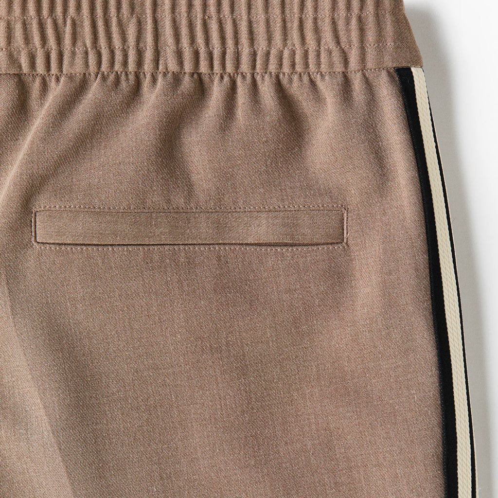 Close-up of a pair of taupe-brown track pants showing the side pocket, elastic waistband, and a black and white side stripe detail.