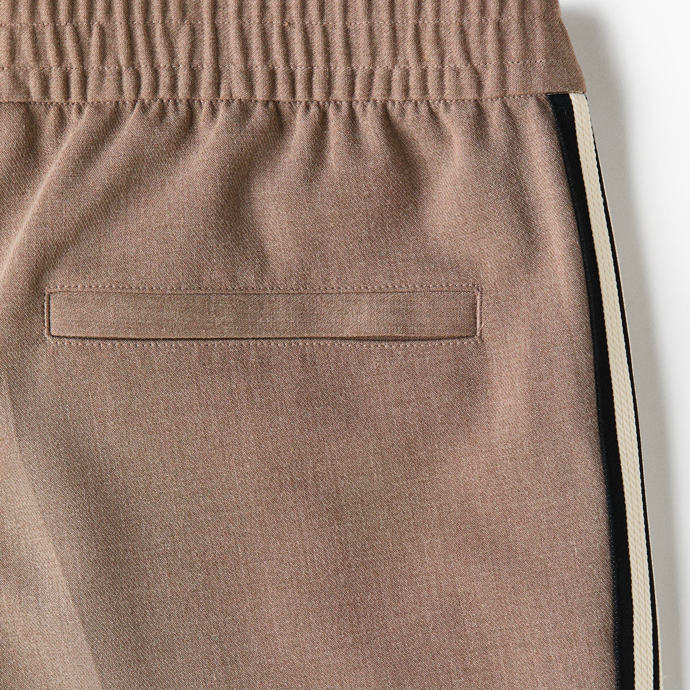 Close-up of a pair of taupe-brown track pants showing the side pocket, elastic waistband, and a black and white side stripe detail.