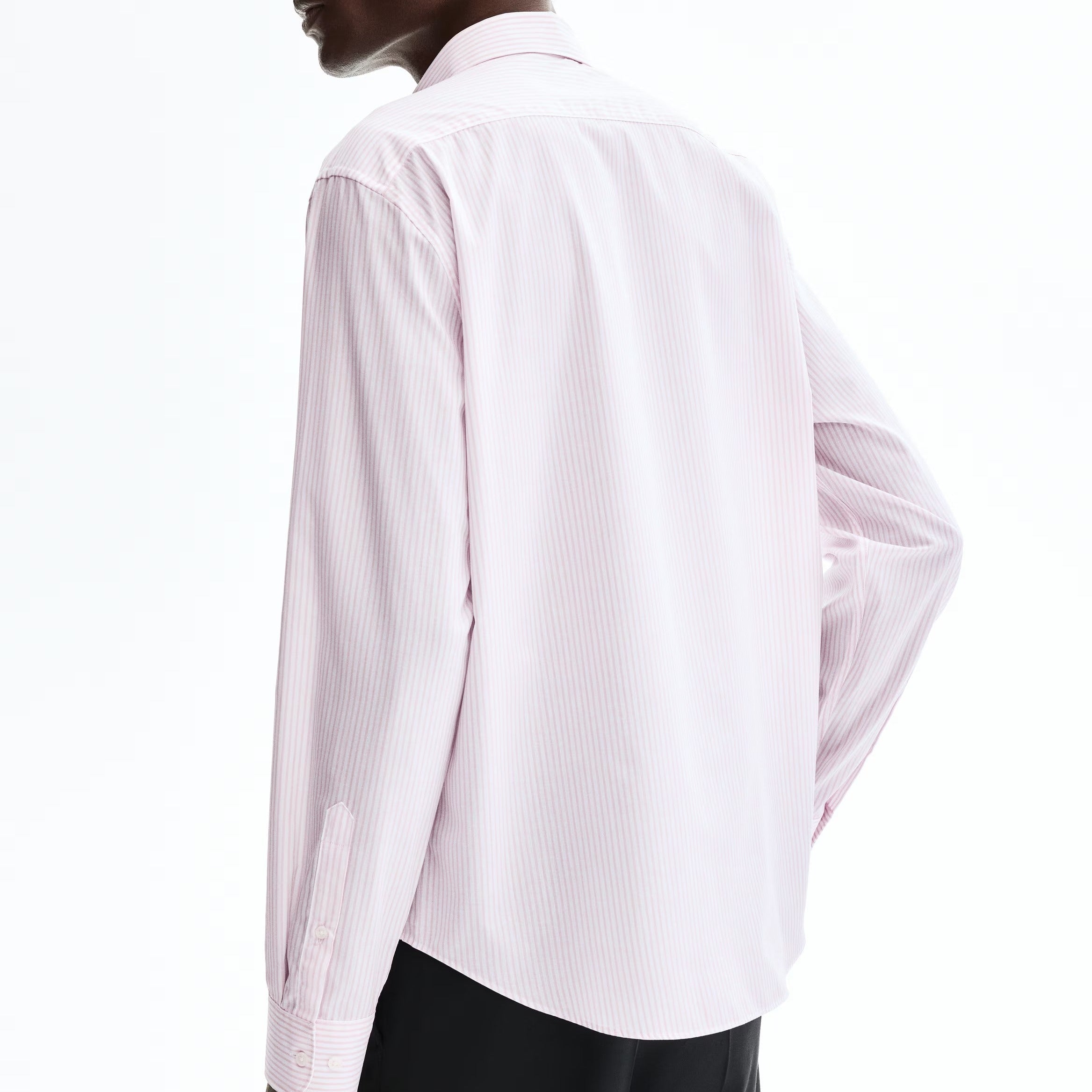 A man wearing a light pink long-sleeve button-up shirt and black trousers, shown from a side-back angle against a plain white studio background.