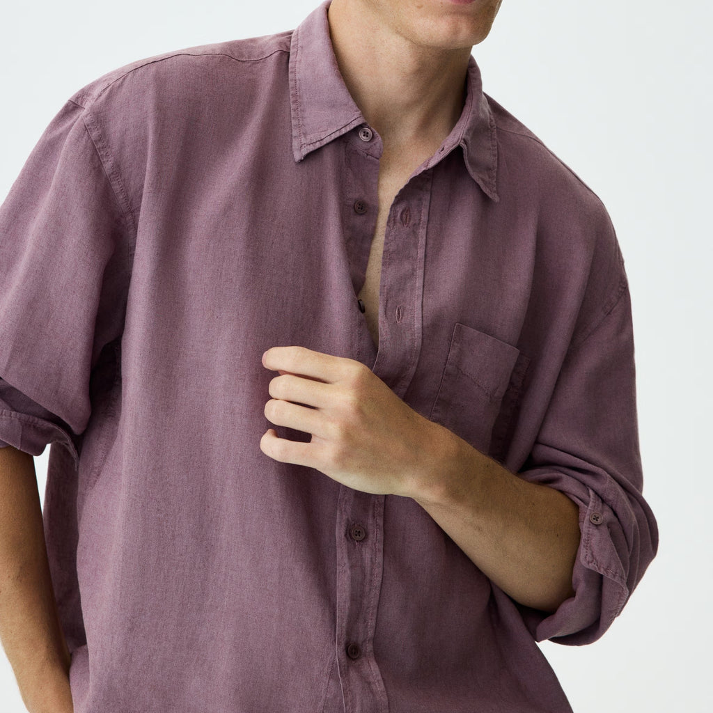 A man wearing a dusty purple, long-sleeve linen button-up shirt with rolled-up sleeves, shown against a plain light gray background.