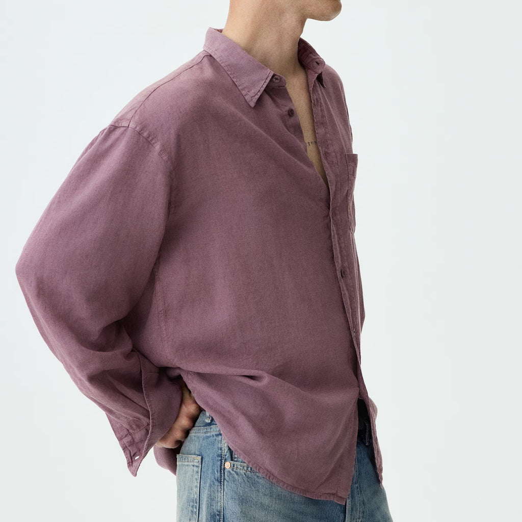 A man wearing an oversized, dusty mauve-purple linen button-down shirt and blue jeans, shown from a side profile against a plain white studio background.
