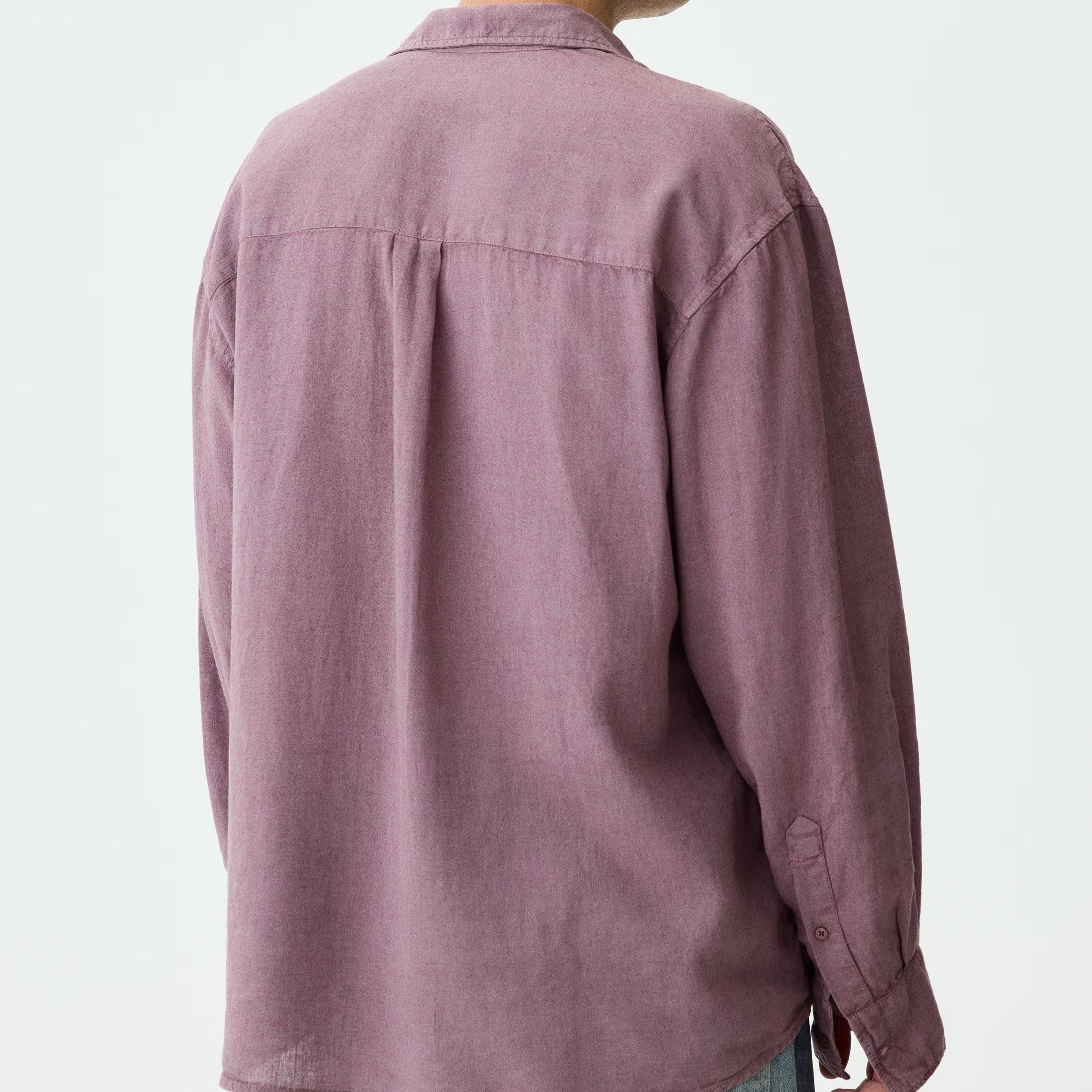 A man shown from the back wearing a dusty mauve-purple long-sleeve linen shirt and light blue jeans against a plain white studio background.