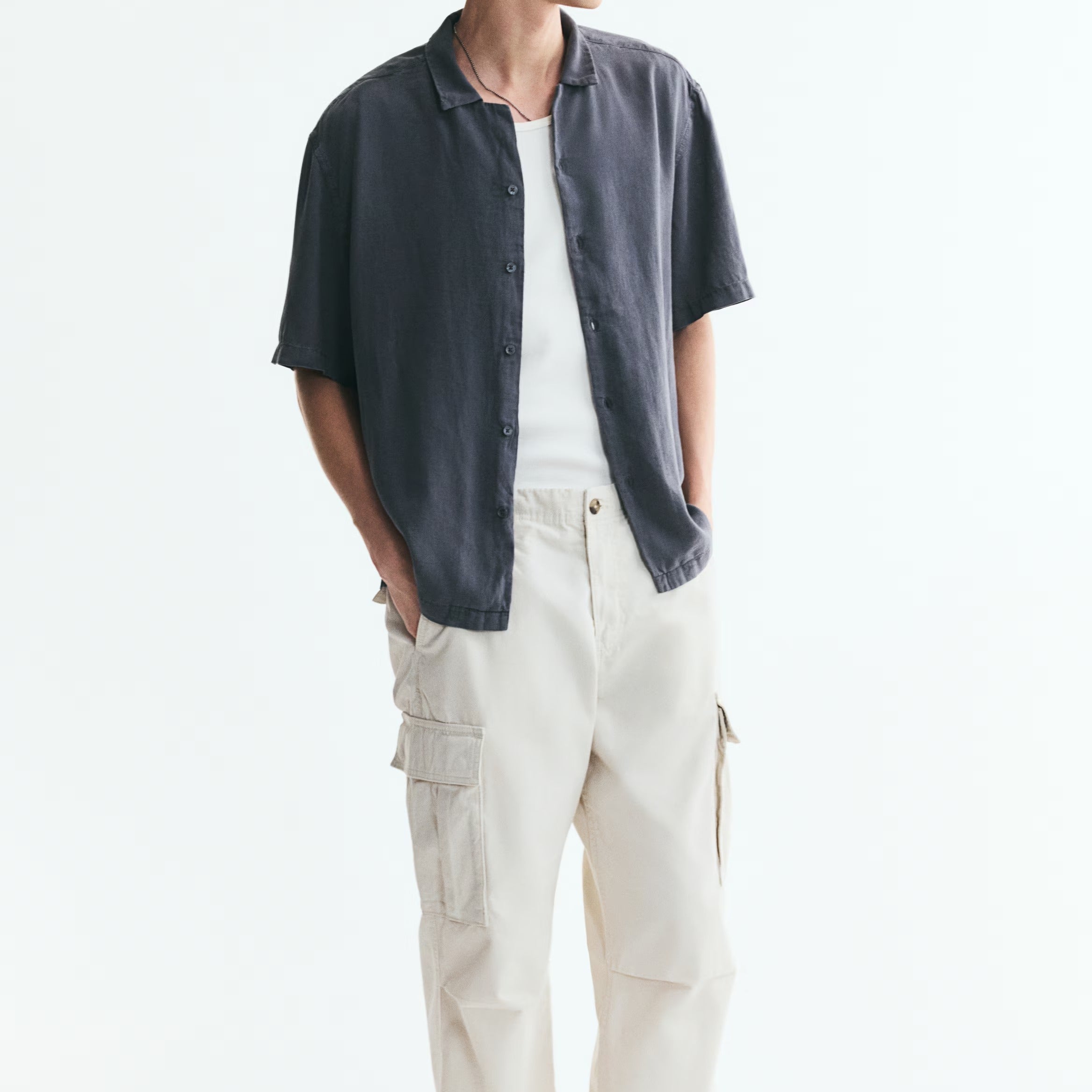 A man wearing a dark navy short-sleeve button-up shirt over a white t-shirt, paired with off-white cargo pants and black slip-on shoes, standing against a plain white background.