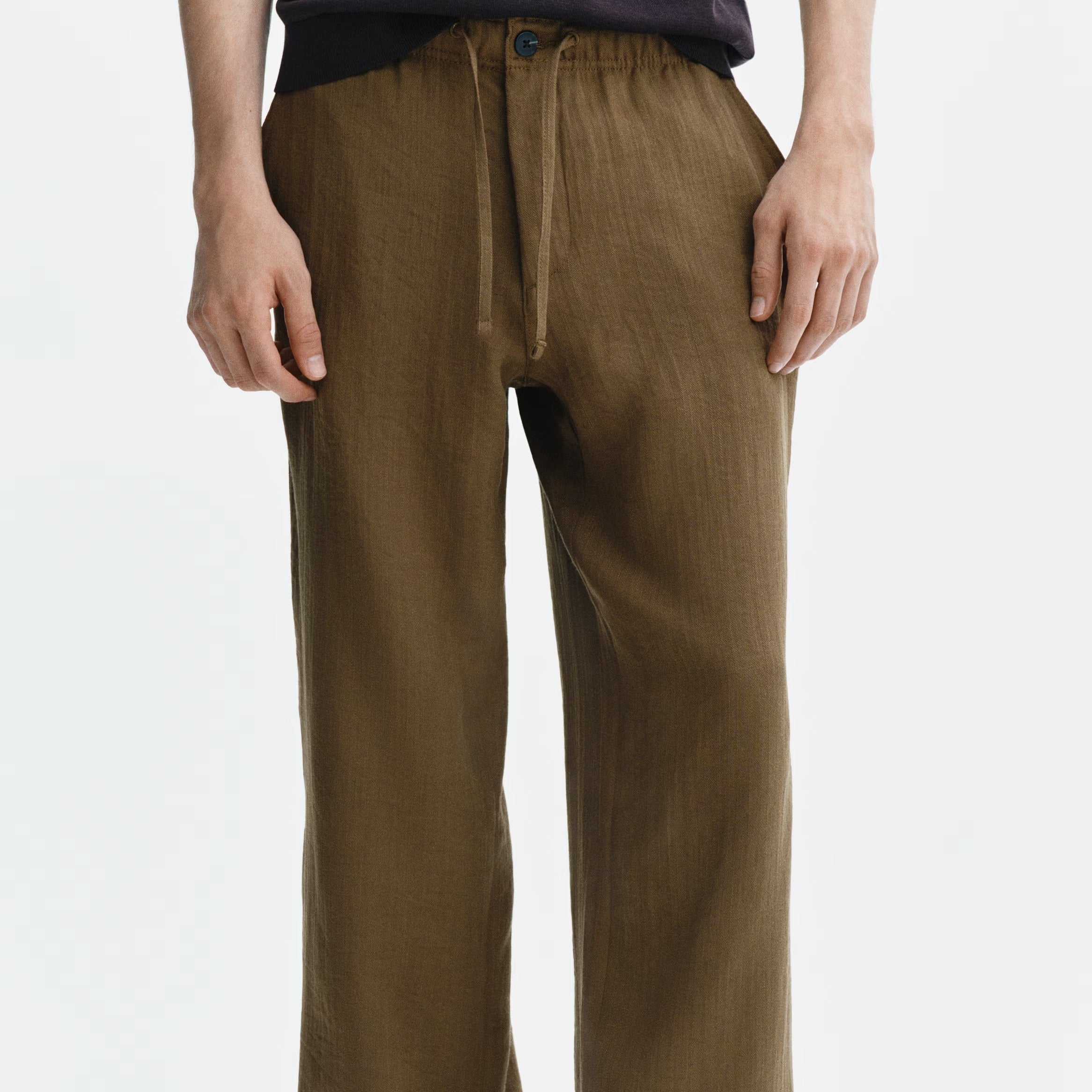 A person wearing loose brown drawstring pants with a dark top and black slide sandals, shown from the waist down against a plain light background.