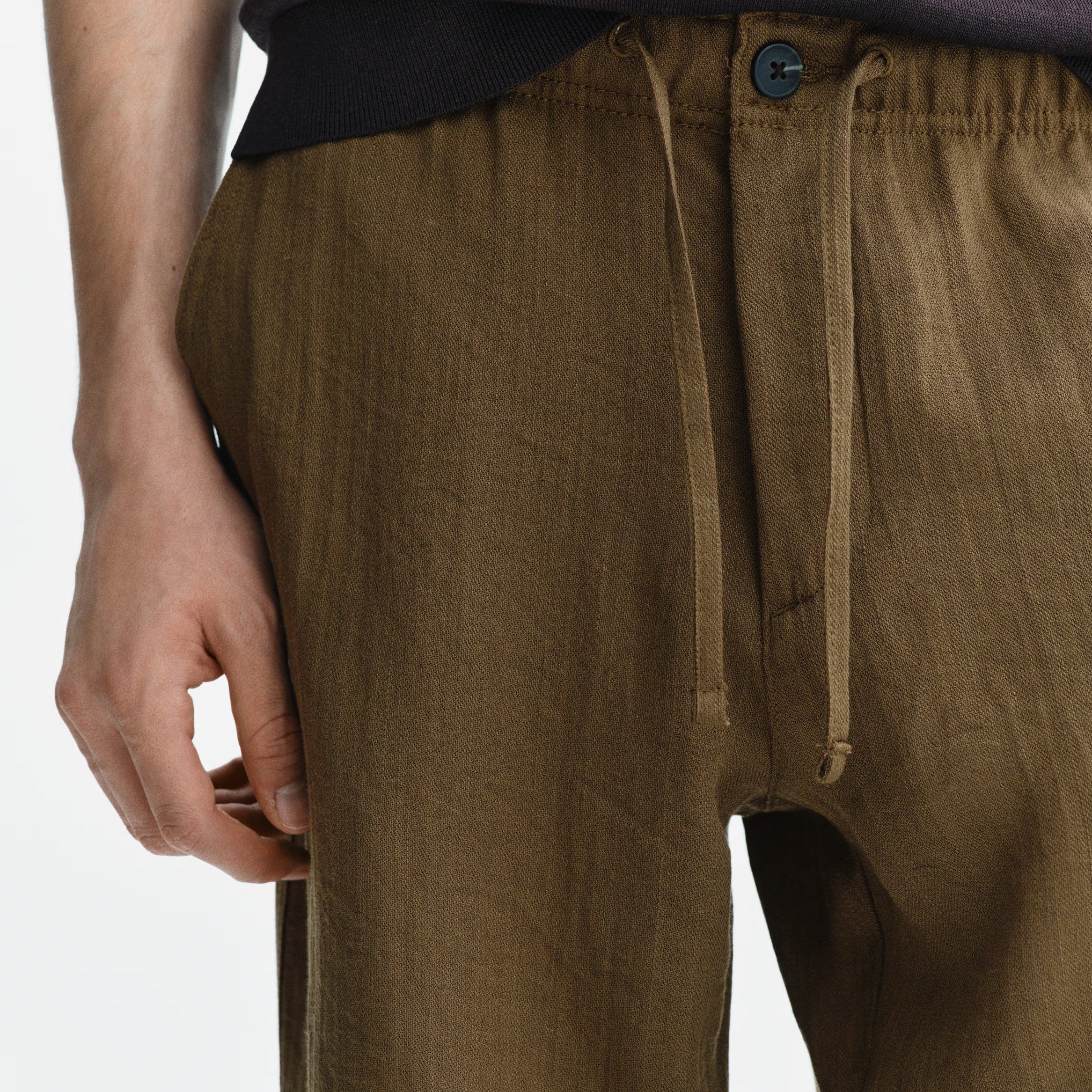 Close-up of a person wearing olive brown drawstring trousers with a dark top, photographed against a plain light background.