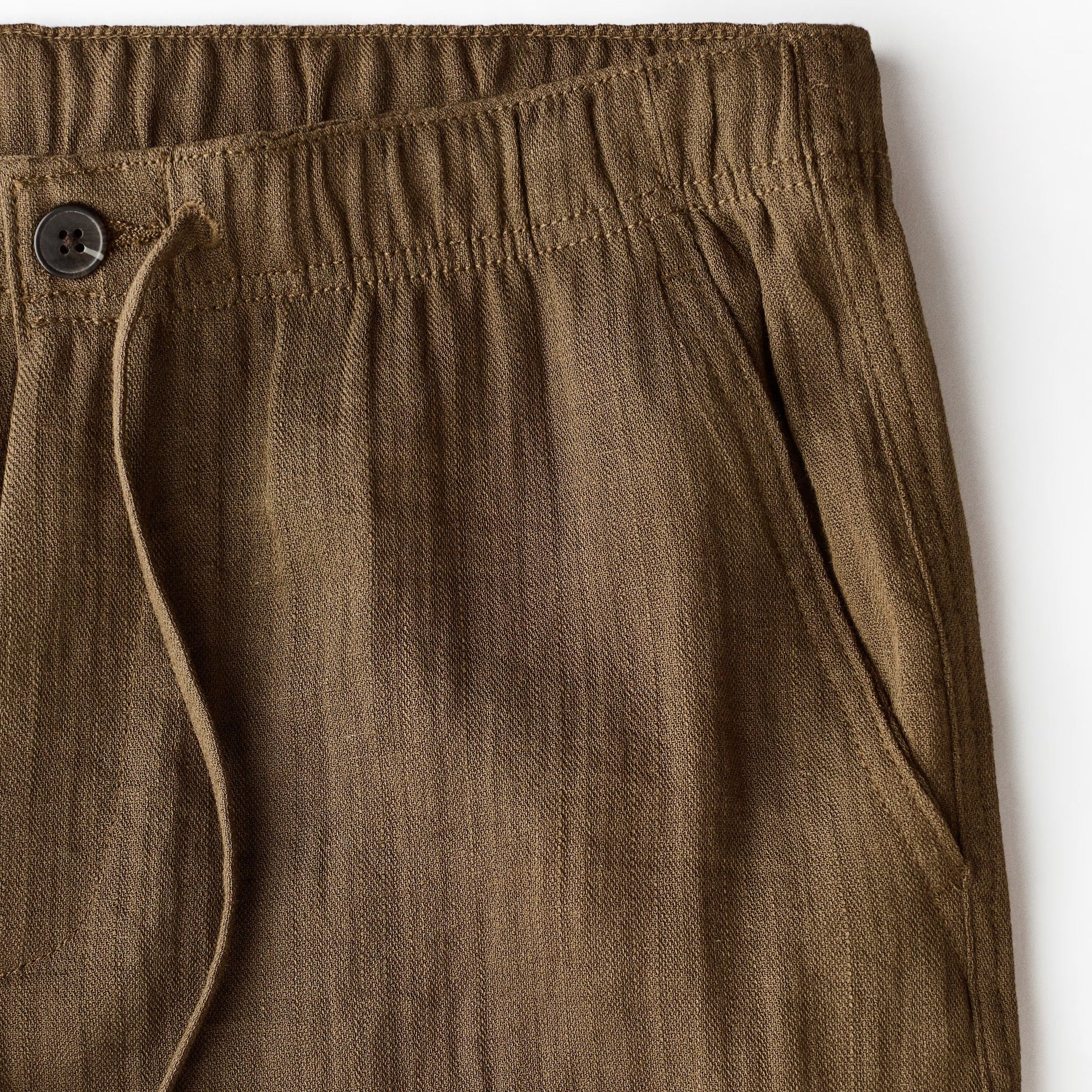 Close-up of olive brown textured trousers showing the elastic waistband, drawstring, and side pocket seam against a white background.