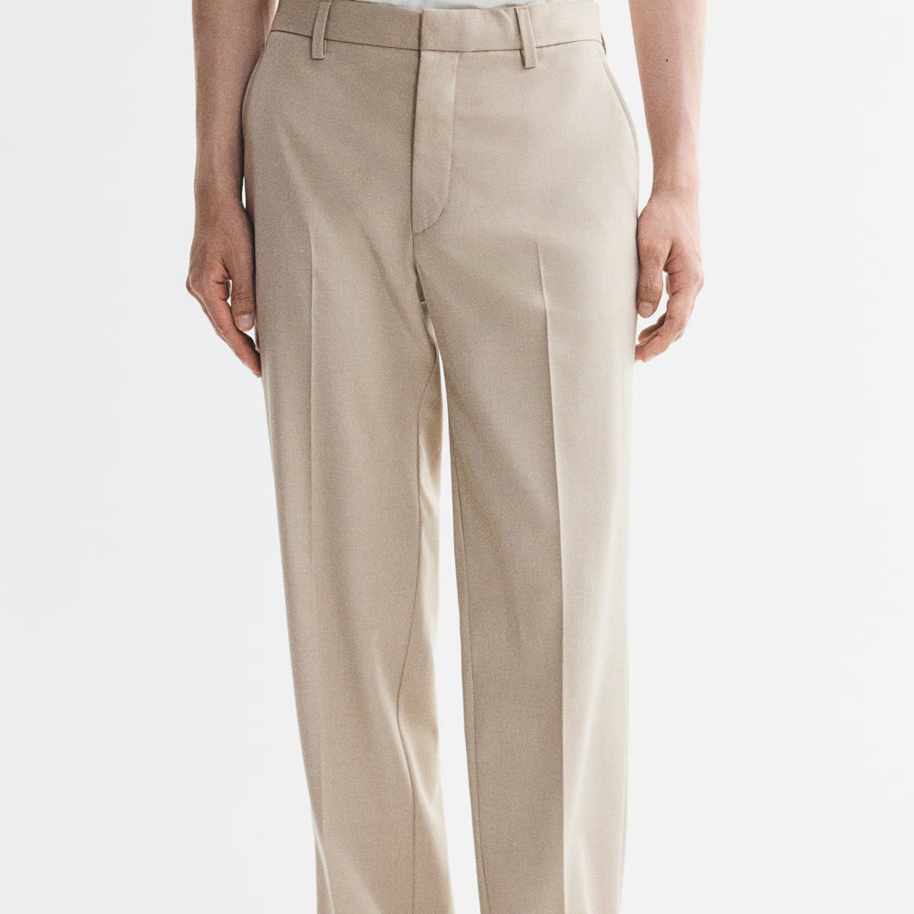 A person wearing light beige straight-leg dress pants with a white top and black flip-flop sandals, shown from the waist down against a plain white studio background.