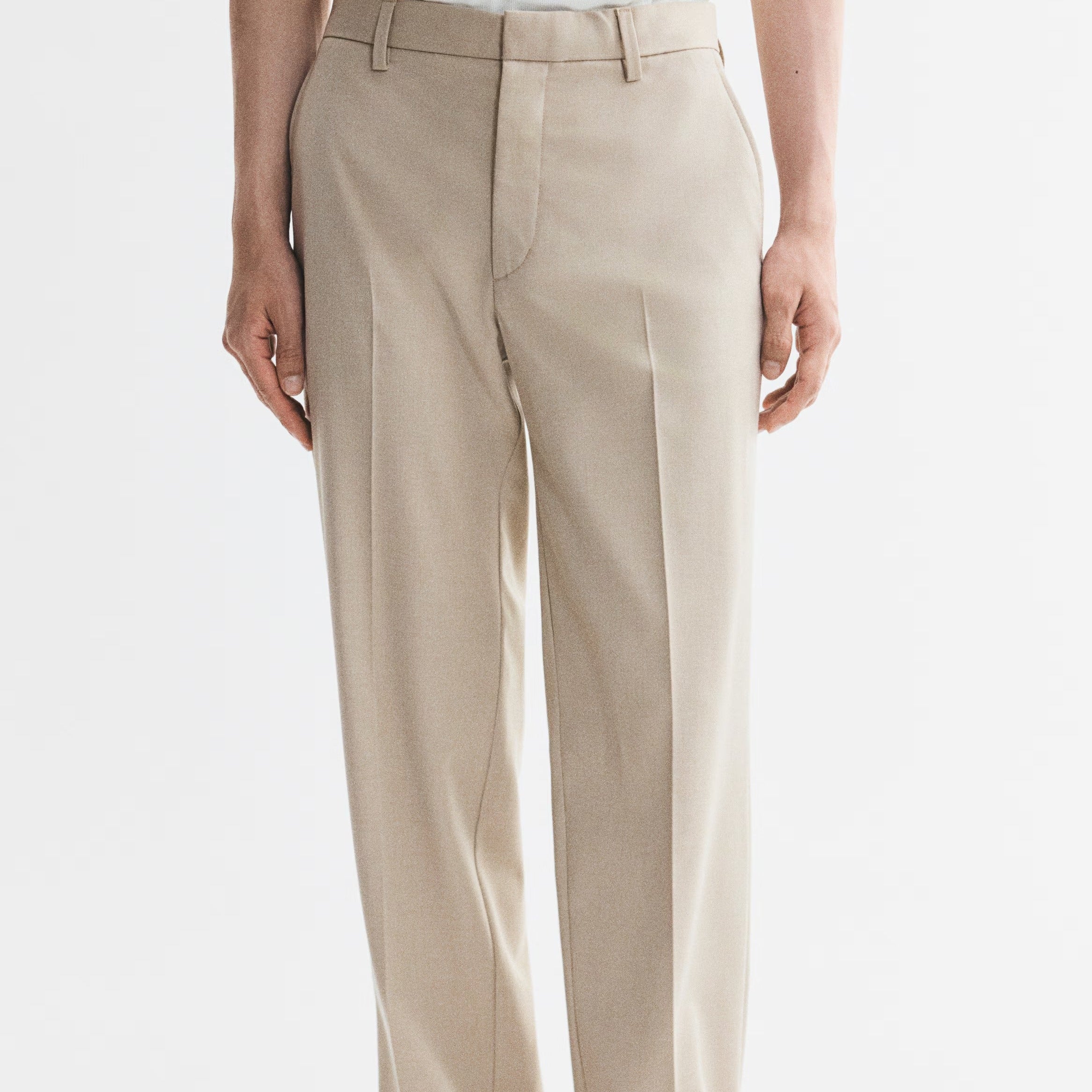 A person wearing light beige straight-leg dress pants with a white top and black flip-flop sandals, shown from the waist down against a plain white studio background.
