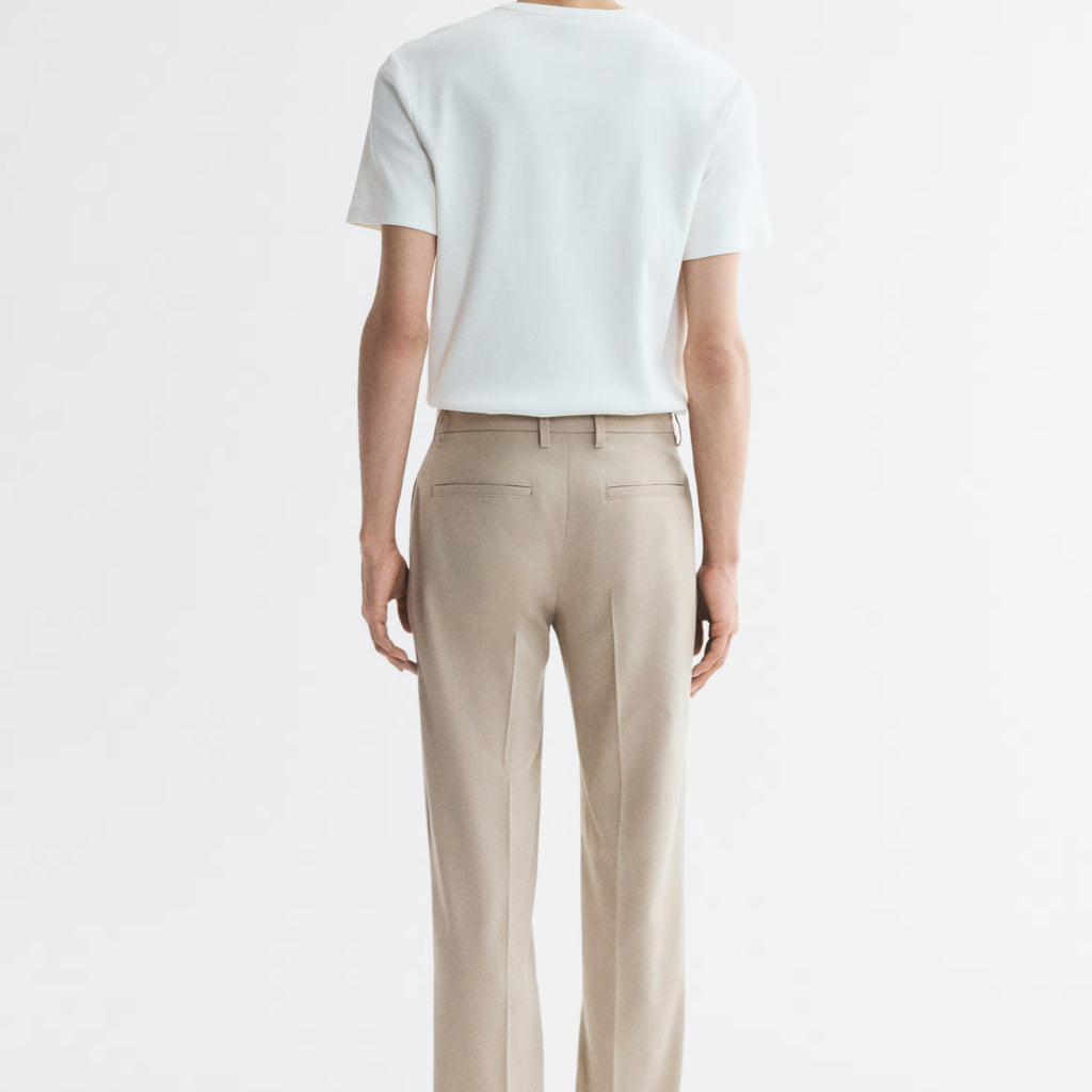 A man viewed from behind wearing a white short-sleeve t-shirt and beige straight-leg trousers, standing against a plain white studio background.