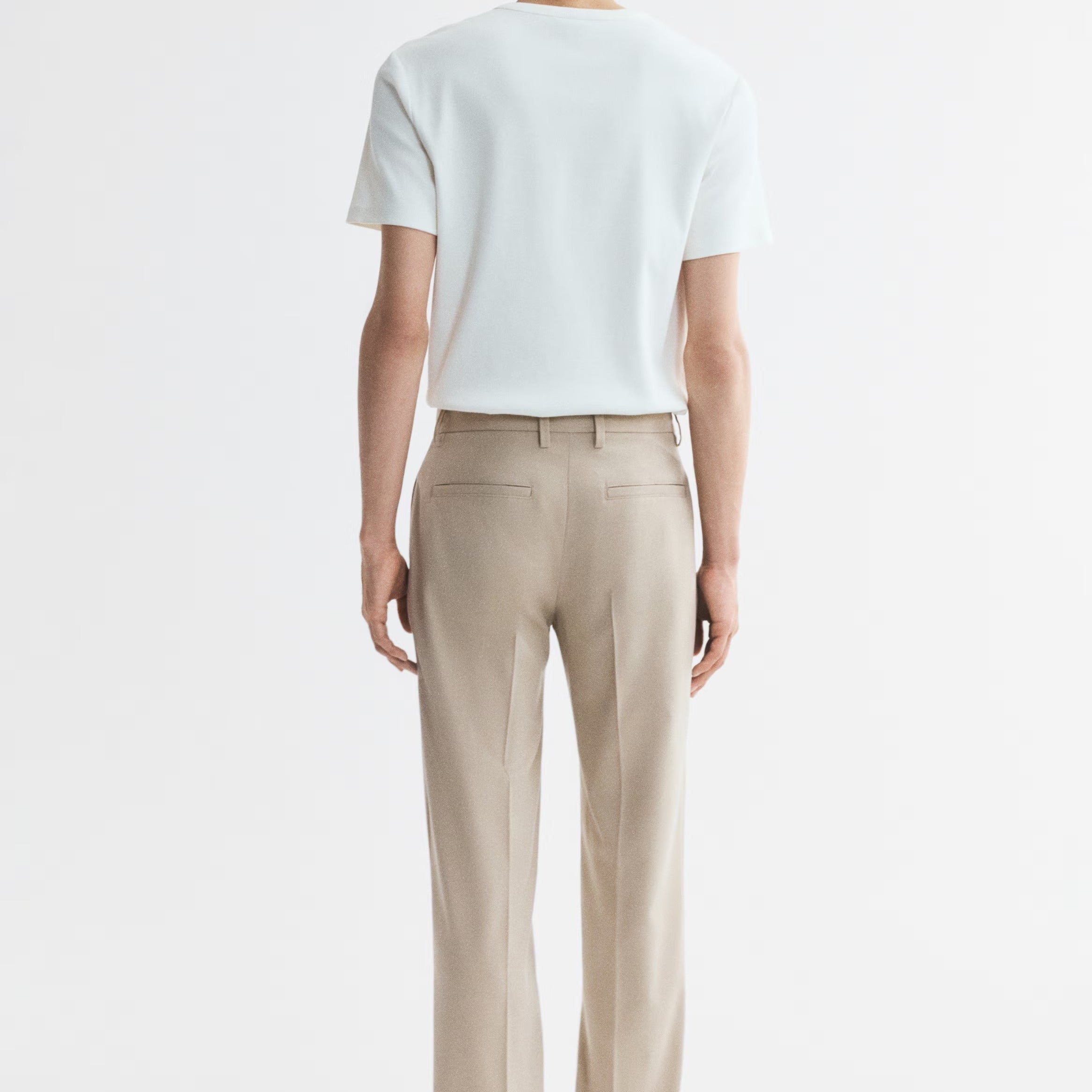 A man viewed from behind wearing a white short-sleeve t-shirt and beige straight-leg trousers, standing against a plain white studio background.