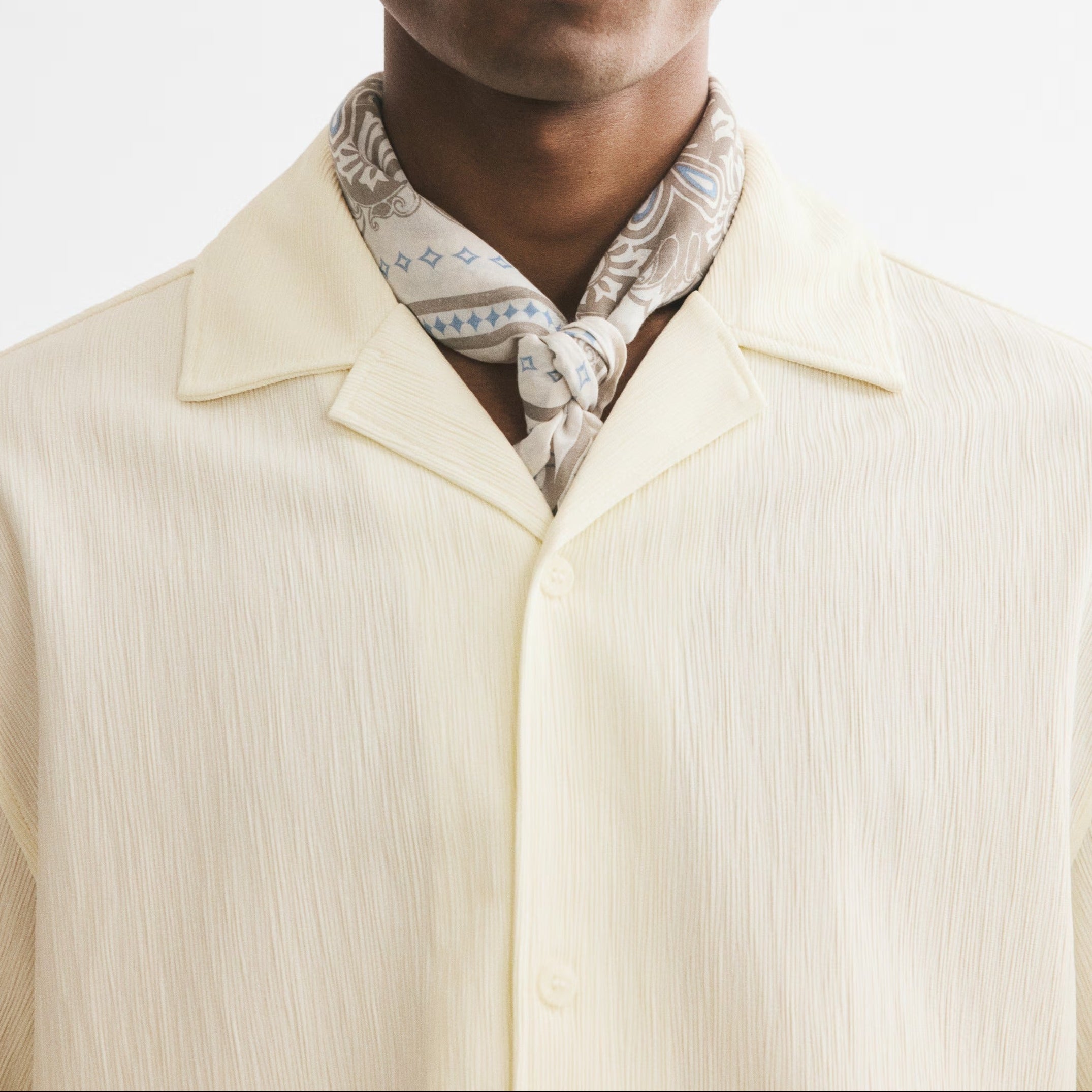 A man wearing thick black square sunglasses, a cream-colored textured collared shirt, and a patterned cream and gray neck scarf, photographed against a plain white background.