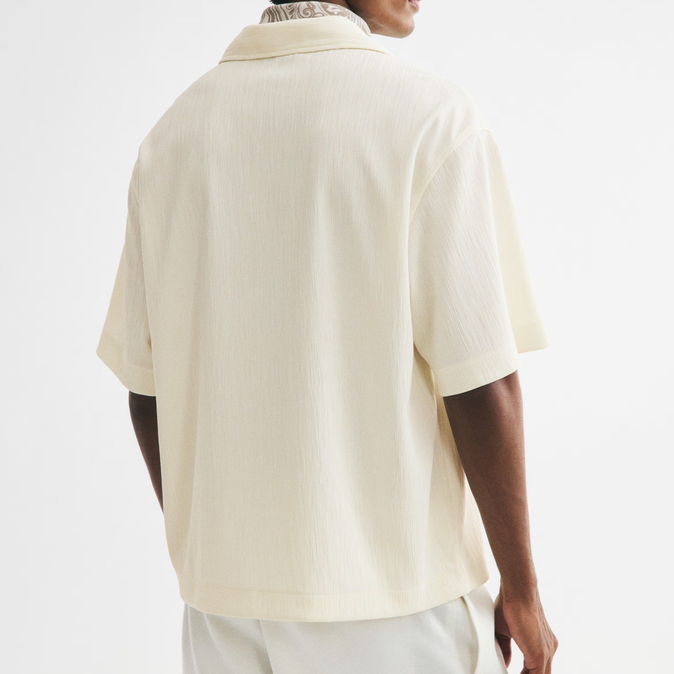 A man shown from the back wearing a cream-colored short-sleeve button-up shirt and matching cream trousers, photographed against a clean white studio background.
