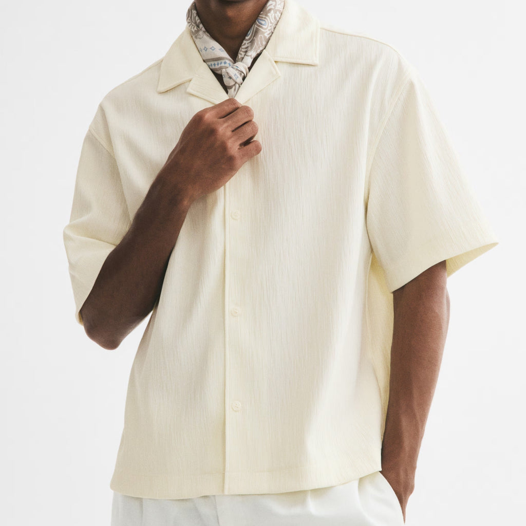 A man wearing a cream-colored short-sleeve button-up shirt, white trousers, a patterned neck scarf, and black sunglasses, posed against a solid white studio background.