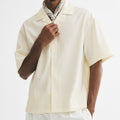A man wearing a cream-colored short-sleeve button-up shirt, white trousers, a patterned neck scarf, and black sunglasses, posed against a solid white studio background.