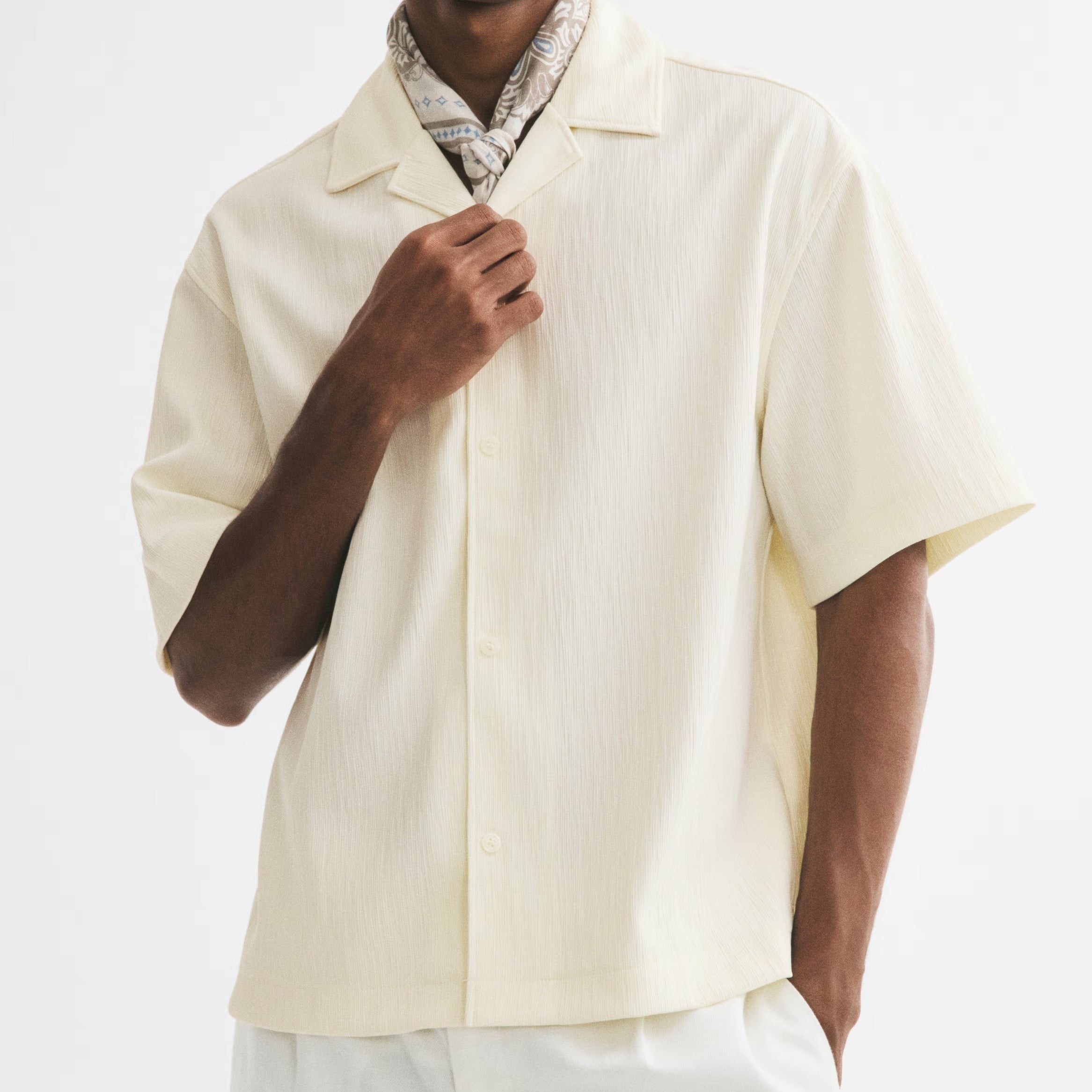 A man wearing a cream-colored short-sleeve button-up shirt, white trousers, a patterned neck scarf, and black sunglasses, posed against a solid white studio background.